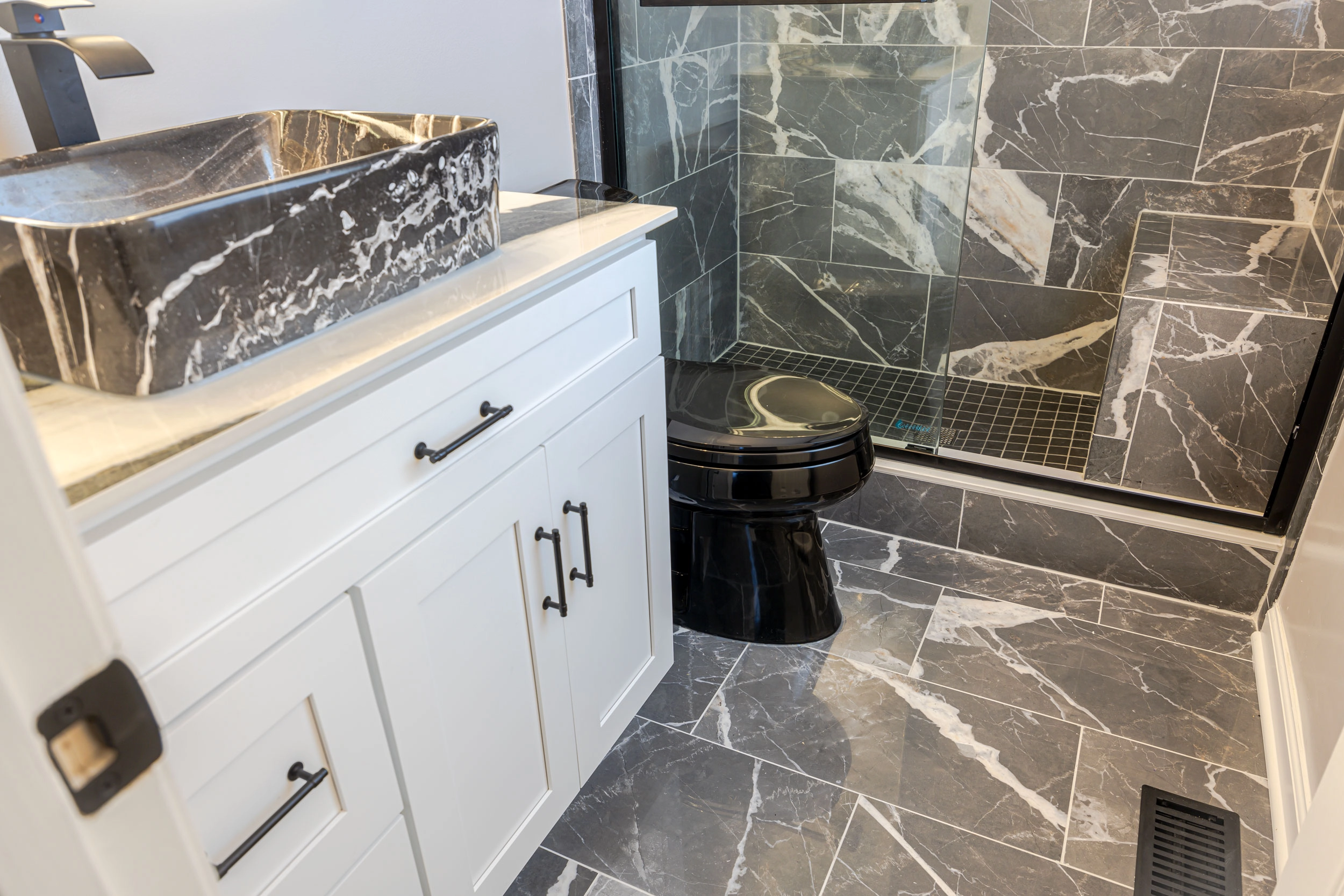 A luxurious bathroom transformation in Bowie, Maryland, by Zion Home Remodeling®. Features black marble-look shower tiles, mosaic floor, sleek white vanity with black vein countertop, vessel sink, black toilet, and oil-rubbed hardware.