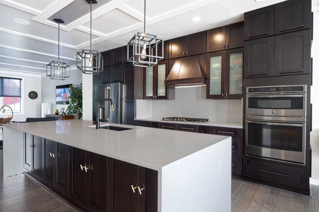 Upper level corner view of an entire remodeled kitchen in Upper Marlboro, Maryland, US, showcasing a large island with Calacatta Unique Gold quartz countertop, Pepper Shaker cabinets, black and gold faucet, three pendant lights, refrigerator with pantries, cooktop area with matching backsplash, and oven cabinet.